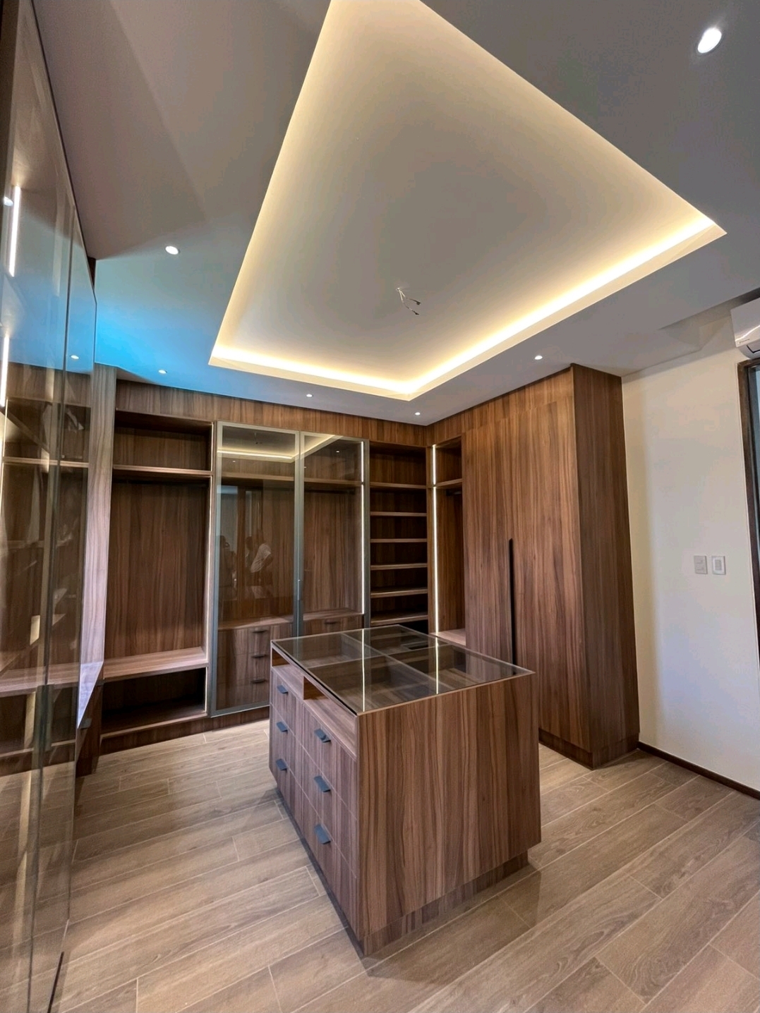 Walk-in closet with island drawers, glass cabinetry and cove lighting